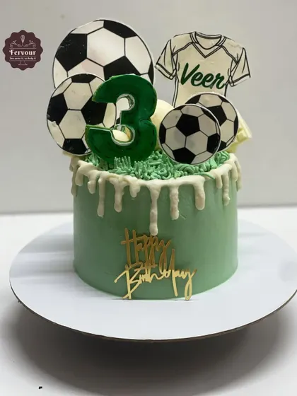 A drip cake with a football theme for a 3rd birthday. The light green cake is topped with edible soccer balls and a jersey, with a white chocolate drip adding a delicious touch.