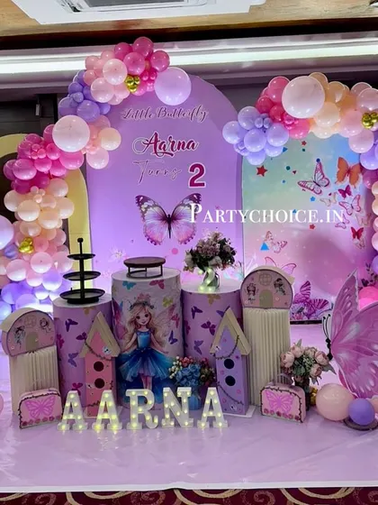 A magical butterfly theme for a second birthday. This elaborate setup features large butterfly wing props, themed pedestals, and a pastel balloon arch against a purple backdrop.