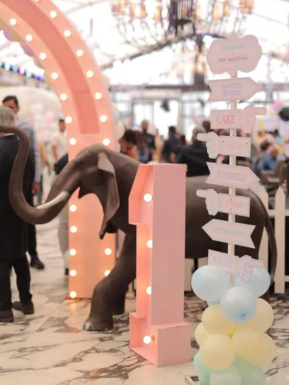 An indoor carnival entrance featuring a light-up marquee number '1' and a life-sized elephant. The directional signpost and pastel balloon clusters guide guests to the fun.