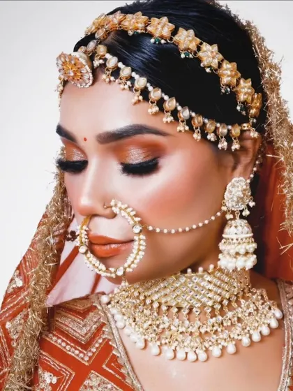 This bride is adorned in a classic gold and pearl jewellery set. The look includes a floral matha patti, a detailed choker, and a traditional nath, perfect for a timeless wedding look.