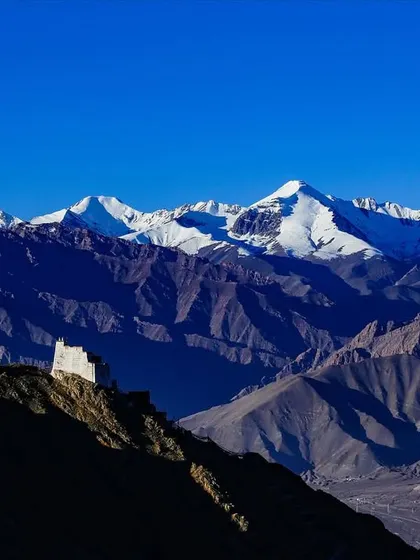 A lone monastery stands on a hilltop, dwarfed by the massive, snow-covered peaks of the Himalayas behind it.