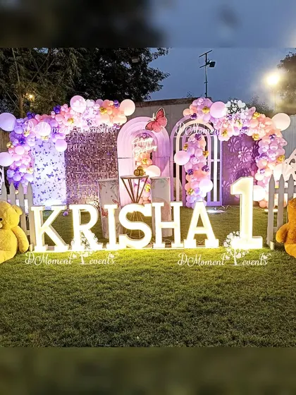 A wide shot of the butterfly-themed lawn party, showing the full stage setup with marquee letters, teddy bears, and a white picket fence.
