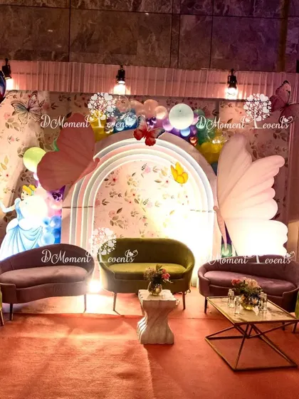 A cozy and elegant seating area designed within the party theme, featuring plush sofas, a floral backdrop, and giant butterfly wing props for a unique photo opportunity.