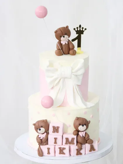 An adorable teddy bear-themed cake for a first birthday. This two-tiered design features a large fondant bow, cute teddy bear figures, and personalized letter blocks spelling out the baby's name.