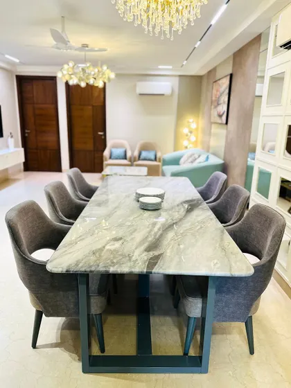 This dining room exudes luxury with a large marble-top table, plush grey upholstered chairs, and two distinct, elegant chandeliers providing ample light.