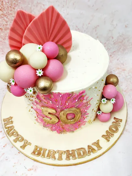 We celebrate a 50th birthday with this vibrant and modern pink boho cake. A bold pink starburst design with a gold "50" is the centerpiece, complemented by pink, white, and gold spheres and dried palm leaves for a chic, bohemian look.