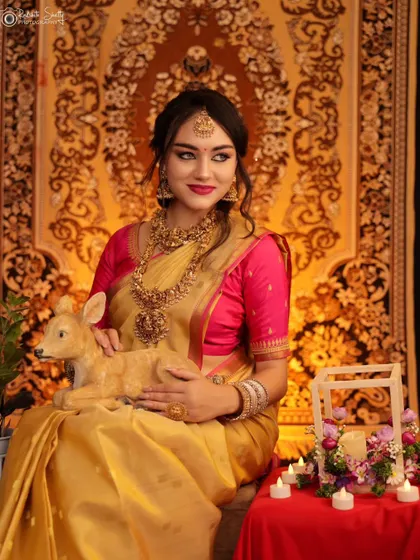 A whimsical and elegant portrait of a model in a golden saree, seated with a small deer figurine. The setup with candles and a traditional rug creates a unique, story-like feel.