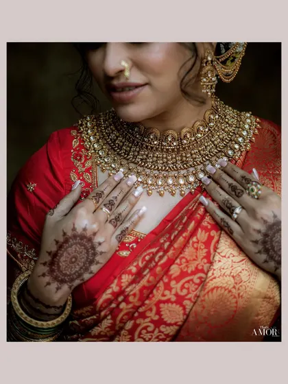 A close-up on the bride's hands and jewelry. This shot highlights the intricate details of her mehndi, rings, and the heavy gold necklace against her red saree.