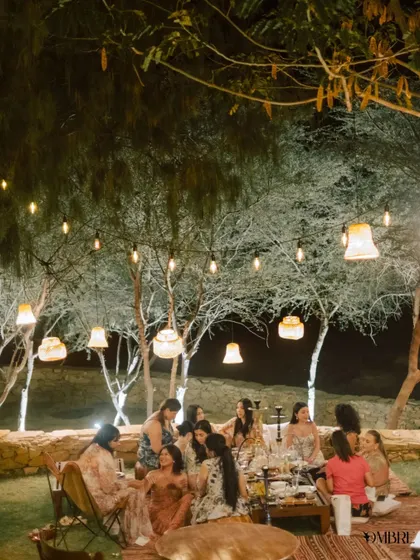 The bride tribe enjoying a relaxed evening outdoors, sitting on rugs under trees strung with lights.