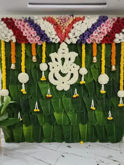 A traditional backdrop for a home ceremony, featuring a Ganesha made of white flowers against a wall of banana leaves. The colorful floral border adds a festive touch.