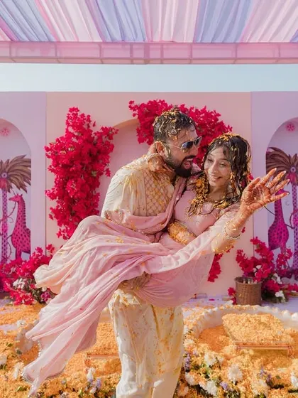 The groom playfully lifts his bride, both covered in turmeric and flowers, a perfect capture of the fun and romance of their Haldi.