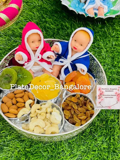 A dry fruit platter for a baby shower celebrating twins. It features two baby dolls in pink and blue robes, surrounded by bowls of cashews, almonds, raisins, and other healthy snacks.