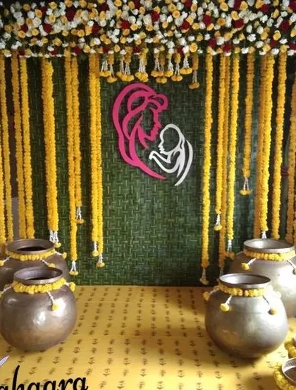 A traditional baby shower or seemantham setup with a backdrop of marigold garlands and brass pots. The mother-and-child motif adds a meaningful touch to the vibrant decoration.