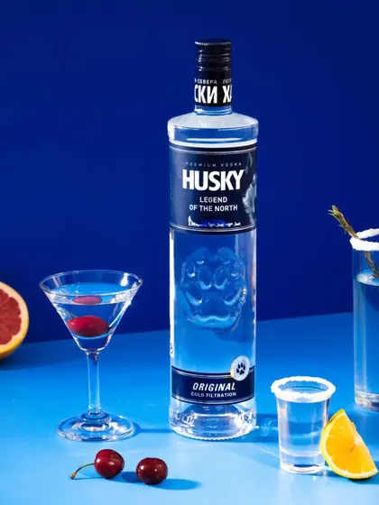 A crisp and clean hero shot for Husky Vodka. The bold blue background and sharp lighting emphasize the bottle's design and the clarity of the liquid, while the cocktail styling adds a touch of class.