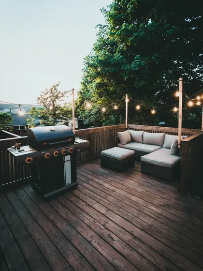 This classic wooden deck is perfect for entertaining, featuring a comfortable L-shaped sofa and a barbecue grill. The string lights add a warm, festive glow for evening get-togethers.