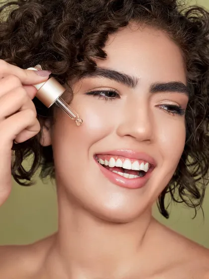 Capturing pure joy in a skincare routine. The model's bright smile while applying a Vitamin C oil from Indulgeo Essentials makes this shot feel energetic and positive. The focus is on the experience of using the product, associating it with happiness and radiant results.