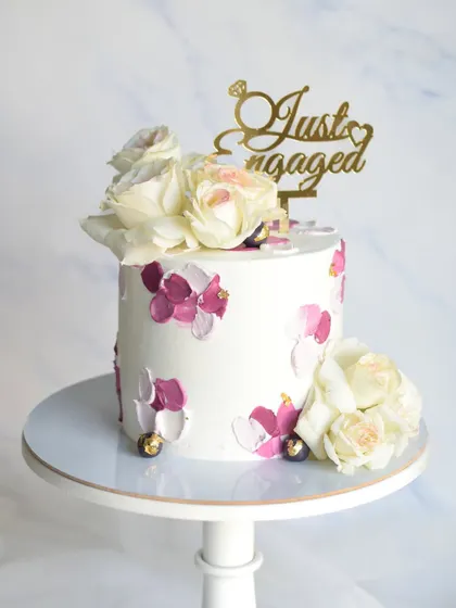 A simple yet beautiful single-tier engagement cake. The design features a palette-knife texture in shades of purple and pink, topped with fresh flowers and a "Just Engaged" topper.