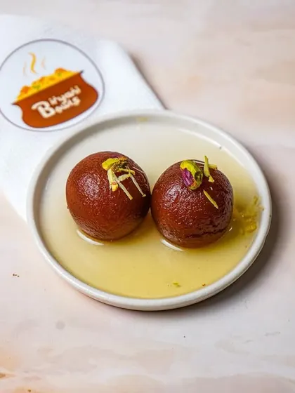 A dark and moody shot of two gulab jamuns in syrup, with the brand's napkin subtly placed. This lighting style adds a touch of elegance and drama to a traditional dessert.