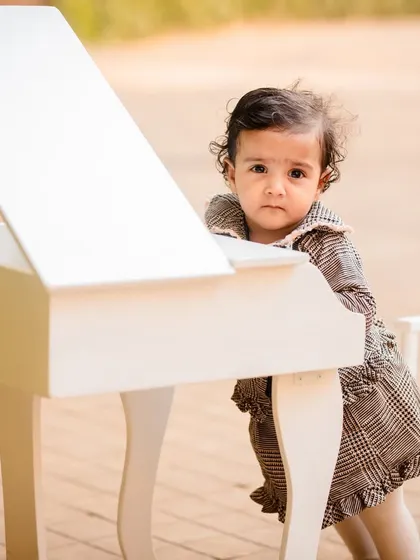 A curious baby girl stands by a miniature piano in a lovely plaid dress during her outdoor photoshoot.
