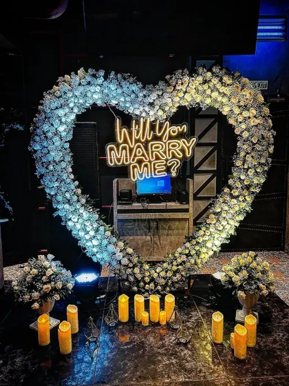 An elegant and dreamy proposal setup featuring a heart-shaped arch of pure white flowers. The glowing candles and central neon sign create a sophisticated and magical ambiance in a darker, intimate setting.