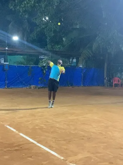 A player tossing the ball for a serve under the bright floodlights. We offer flexible evening slots for practice and matches.