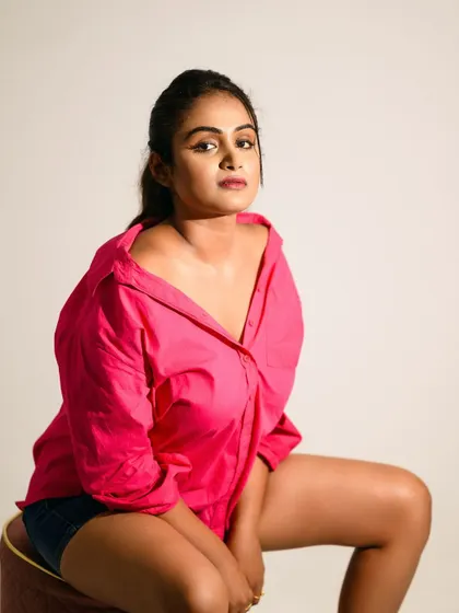 A simple and confident studio portrait with a pink shirt and denim shorts. The direct gaze and relaxed pose create an approachable and modern look.
