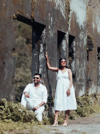 A stylish couple in matching white outfits poses against a rustic, weathered wall, blending modern fashion with a vintage-inspired location.
