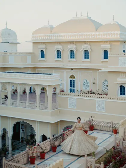 The bride twirls on a palace terrace, her golden lehenga catching the light, with the magnificent building creating a grand backdrop.