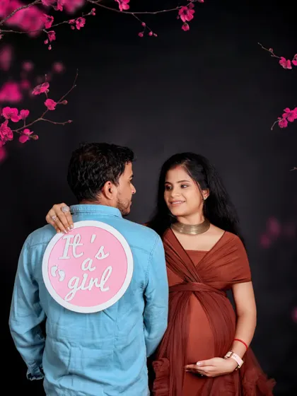 A fun gender reveal shot. The mom-to-be looks lovingly at her partner, who has an "It's a girl" sign playfully placed on his back.