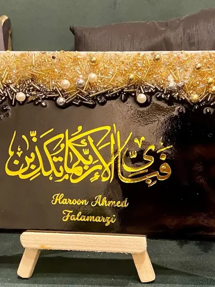 This personalized plaque features the verse "Fabi ayyi ala i rabbikuma tukazziban" in bold yellow calligraphy on a black background. The top is adorned with a textured mix of gold and silver embellishments, and it includes the client's name for a personal touch.