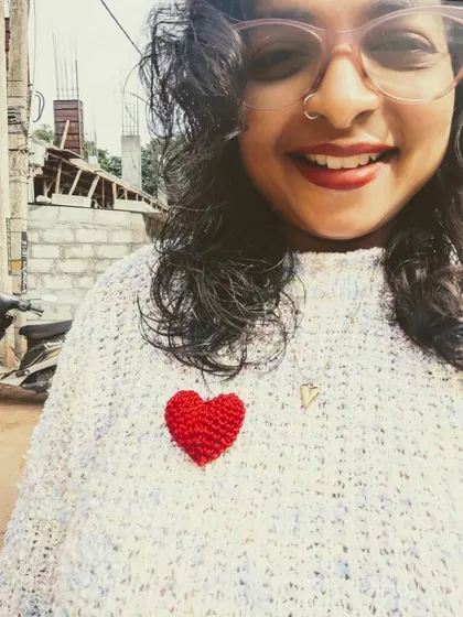 A selfie showing off one of my small, bright red crochet hearts pinned to my sweater. It's a cute little accessory that adds a pop of color and love to any outfit.