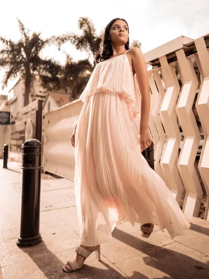 Full-length shot of a model in a pink pleated dress on a sidewalk, with palm trees in the background