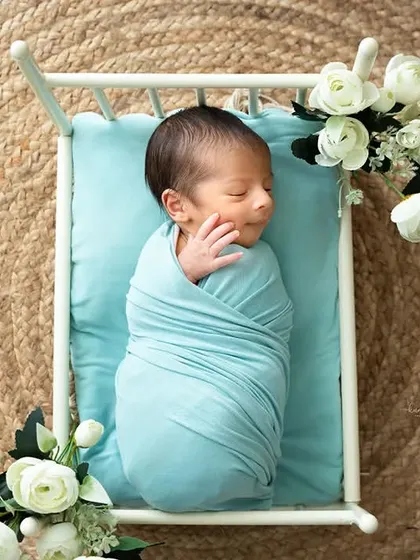 A newborn wrapped in a blue swaddle sleeps in a miniature bed set on a natural jute rug, combining rustic and soft elements.