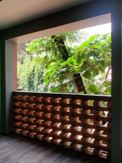 A balcony with a terracotta jali railing, framing a view of the green trees outside and providing ventilation.