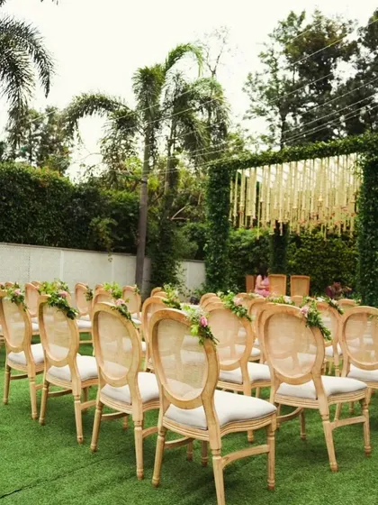 An outdoor ceremony seating arrangement with elegant wooden chairs on a lush green lawn. The aisle is clear, leading to a beautiful mandap covered in greenery.
