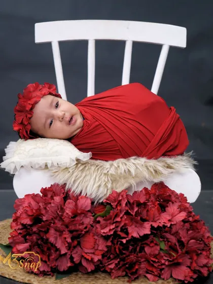 A beautiful baby girl wrapped in red, resting on a miniature white chair surrounded by flowers. The contrast of the red against the dark background makes the image pop.