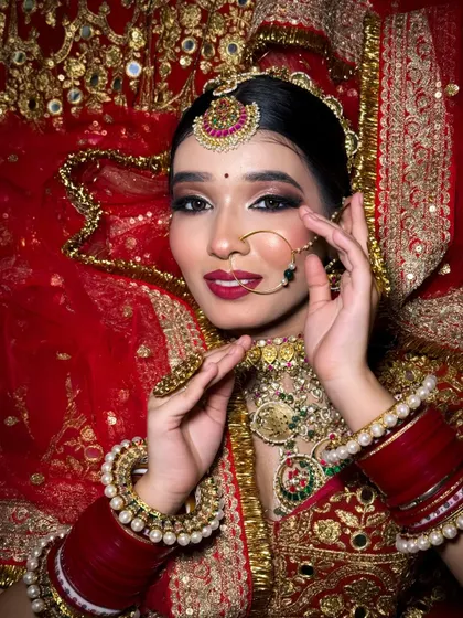 A classic bridal portrait. The bride is looking directly at the camera, showcasing her stunning eye makeup, nose ring, and traditional red attire.
