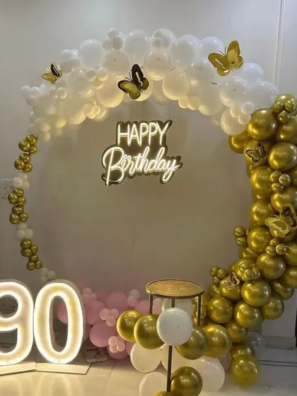 A grand 90th birthday celebration with a white and gold circular balloon arch, a '90' marquee light, and butterfly accents.