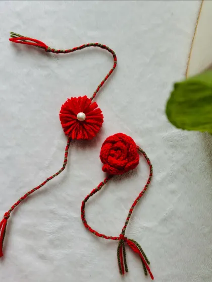 Two of my handmade rakhis in a beautiful, vibrant red. One is a pom-pom flower with a pearl, and the other is a delicate knitted rose. They are a special way to share a bond.