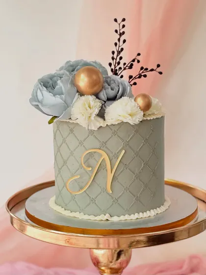 An elegant grey cake with a quilted pattern and a gold monogram. The cake is topped with dusty blue silk flowers, white carnations, and gold spheres.