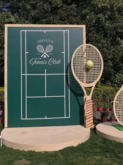 A sophisticated Tennis Club theme for a first birthday. The main photo backdrop features a custom tennis court design, oversized rackets, and a grass-covered number '1'.