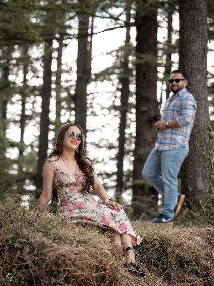 A relaxed and happy portrait of the couple in the woods, with her sitting on a grassy patch while he leans against a tree.