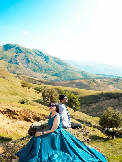 A breathtaking wide shot of a couple sitting on a rock, with the woman's blue gown spread out, overlooking a vast expanse of green hills.