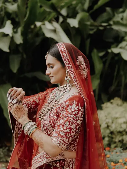 A side profile of the bride, showing her adjusting her bangles. Her makeup is soft and elegant, a perfect match for her red Pheras lehenga.