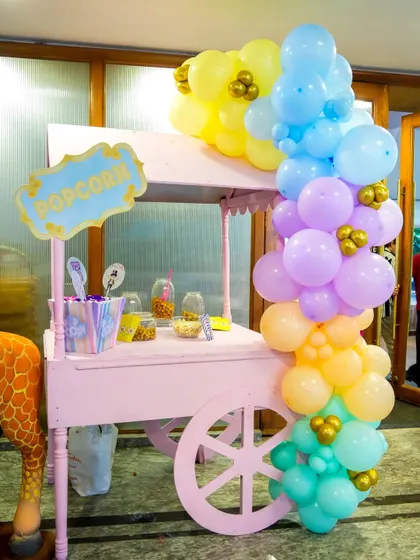 A pink popcorn cart adorned with pastel rainbow balloons and gold accents for a whimsical carnival party.
