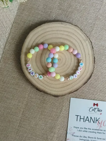Matching pastel name bracelets for Siana and Saina, displayed on a wooden slice with our thank you card.