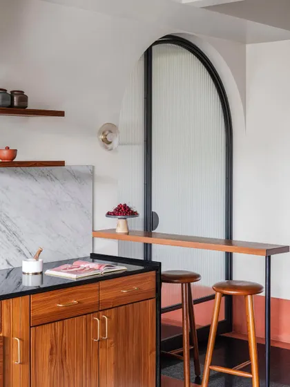 A detail of the kitchen, showing the combination of rich timber cabinetry, black granite, white marble, and brass handles. A small bar counter provides a casual spot for a morning coffee.