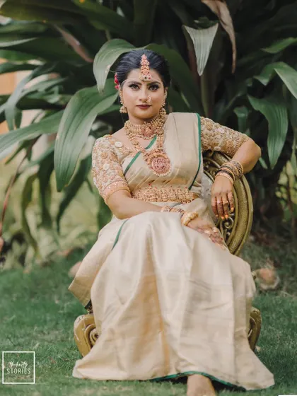 A full-length portrait of a bride in a cream and gold saree, seated gracefully outdoors, looking regal and poised.