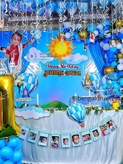 For this first birthday, I designed a sky and hot-air balloon theme. The decor features a custom backdrop, blue and white balloon clouds, a photo banner showing the baby's first year, and a large '1' foil balloon.