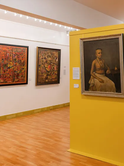 A view of the gallery featuring a portrait by Mangalabhai Thampuratty on a vibrant yellow wall, alongside abstract works by Achutan Kudallur. The layout is designed to spark dialogue between different artistic eras.